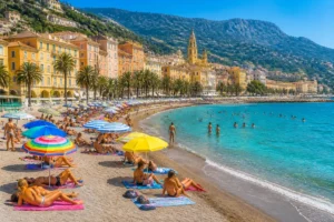 Plage de Menton en été, baigneurs, mer turquoise, façades colorées et montagnes verdoyantes en arrière-plan